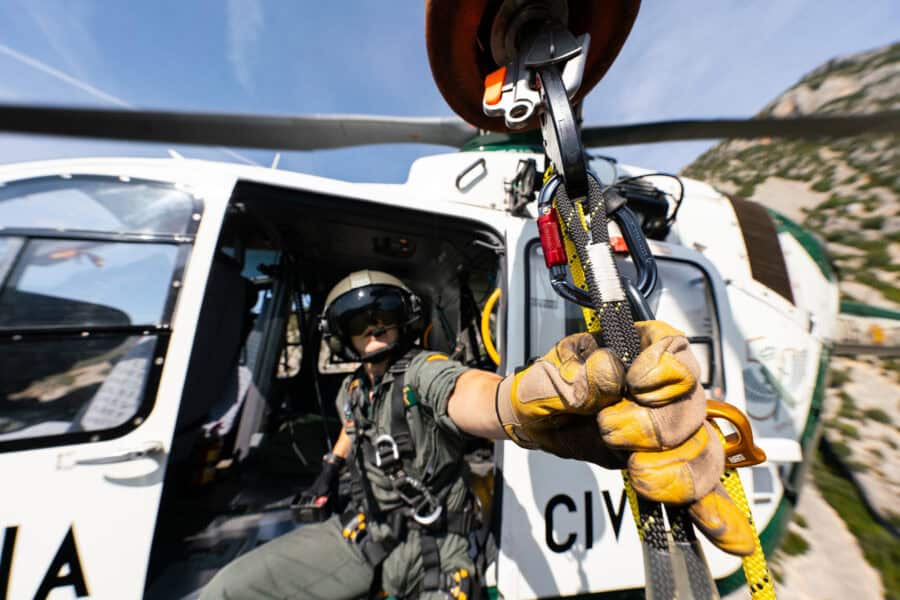 An onsite safety professional riding in a white helicopter reaches out for a safety line with a gloved hand. It is a clear sunny day and there is a mountain in the background.