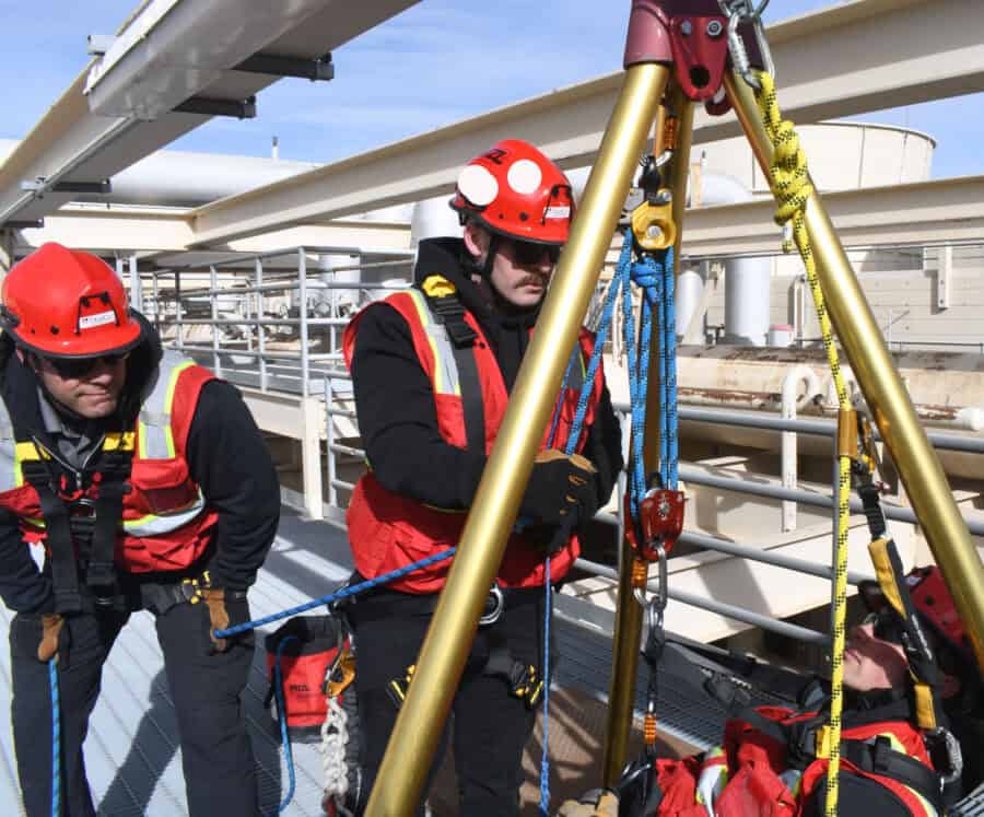 Three workers in safety gear conduct tactical rescue training, operating lifting equipment on an industrial platform with a harnessed person.
