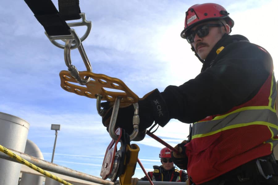 A worker in safety gear, including helmet and sunglasses, adjusts climbing equipment on a rooftop while a fire rescue member is visible in the background.